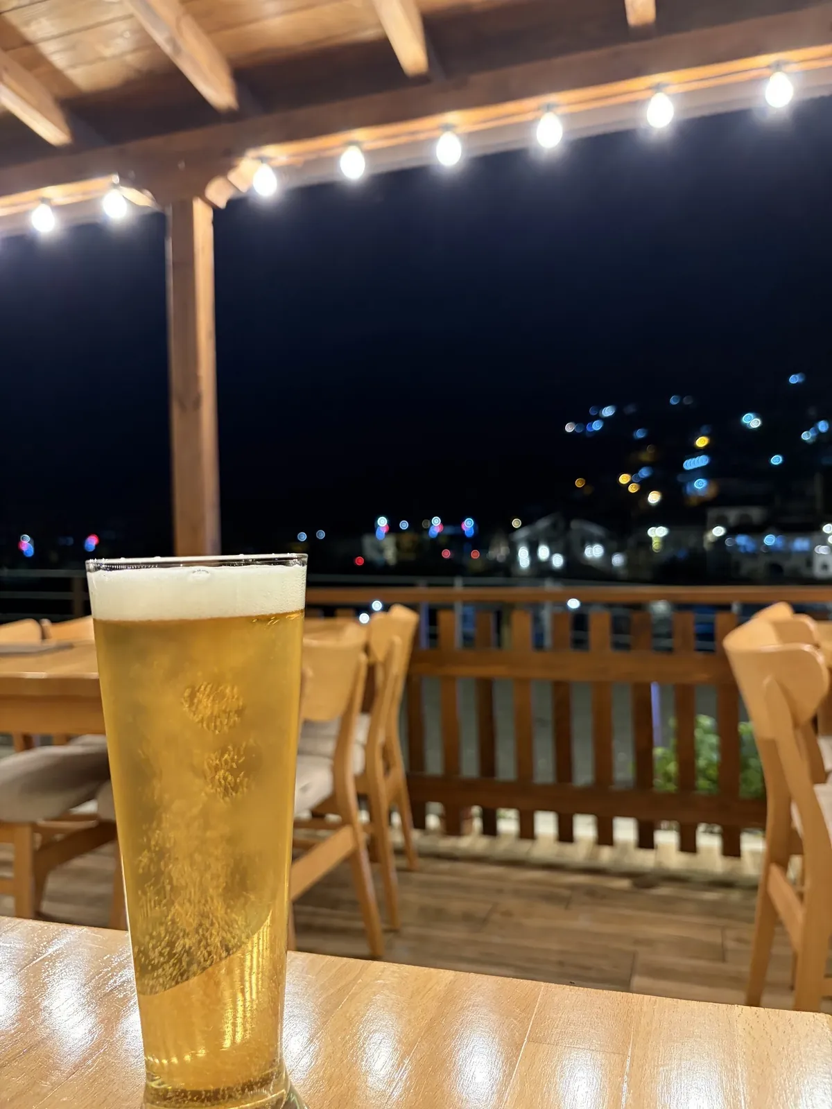 A glass of beer on the terrace at night, with the lights of Berat behind