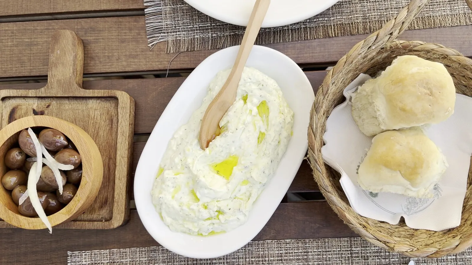 Olives in a wooden bowl, a white spread with olive oil and bread rolls in a basket