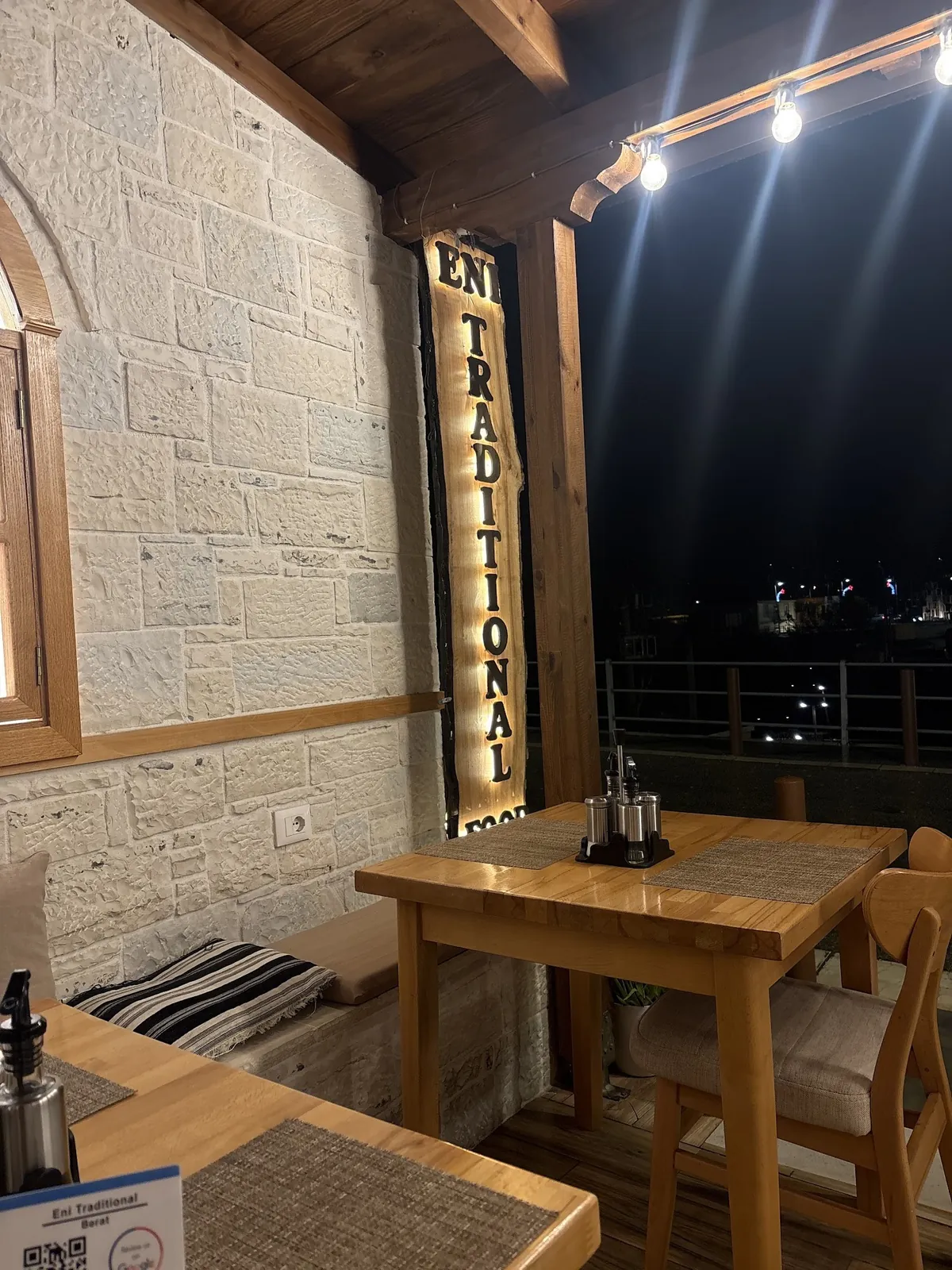 A corner of the terrace at night, with the lit Eni Traditional Food sign and a wooden table